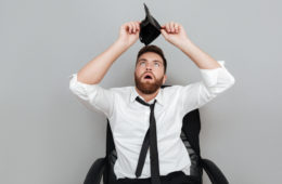 Frustrated surprised bearded man in white shirt showing empty wallet while sitting in chair isolated over gray background